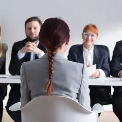Kobieta w szarym żakiecie siedzi przed komisją rekrutacyjną. To jej druga rozmowa kwalifikacyjna, a uśmiechnięci pracodawcy wydają się zadowoleni.