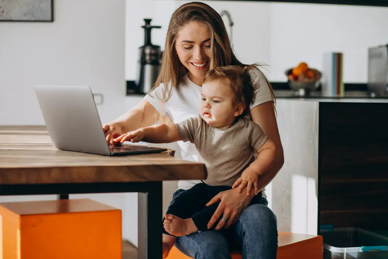Uśmiechnięta mama z dzieckiem na kolanach pracuje przy laptopie. To przykład udanej familijnej pracy, gdzie obowiązki łączą się z opieką nad maluchem.